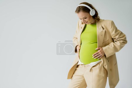 Téléchargez les photos : Femme enceinte élégante dans les écouteurs toucher le ventre isolé sur gris - en image libre de droit