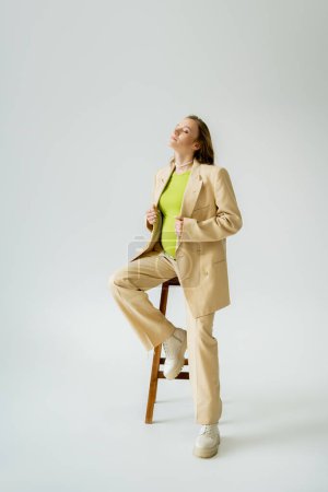 Modische Schwangere im Anzug posiert in der Nähe von Stuhl auf grauem Hintergrund 