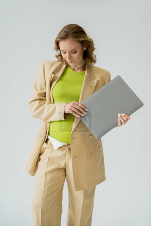 Stylische schwangere Freiberuflerin im beigen Anzug hält Laptop isoliert auf grau 