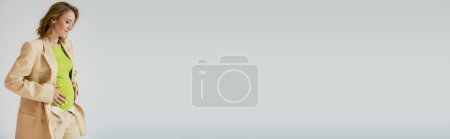 Photo for Young pregnant woman in suit and green blouse touching belly isolated on grey, banner - Royalty Free Image
