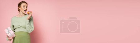Foto de Mujer embarazada con boca abierta sosteniendo rosquilla y caja de regalo sobre fondo rosa, pancarta - Imagen libre de derechos