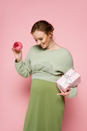 Happy pregnant woman in green outfit holding donut and gift while looking at belly on pink background 