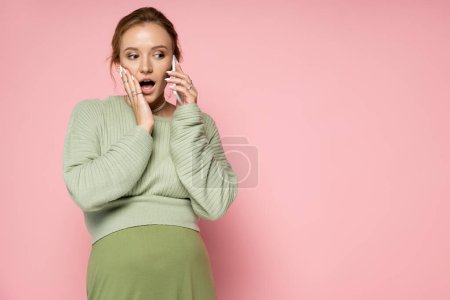 Téléchargez les photos : Choqué femme enceinte en tenue verte parlant sur téléphone portable sur fond rose - en image libre de droit