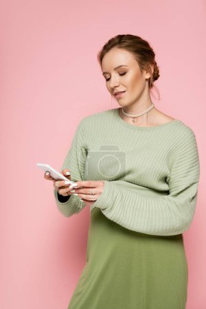 Téléchargez les photos : Femme enceinte élégante en tenue verte en utilisant un téléphone mobile sur fond rose - en image libre de droit