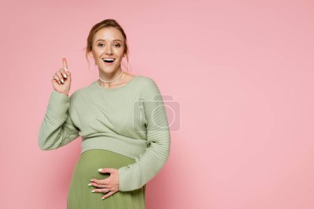 Photo for Excited pregnant woman in green outfit pointing with finger isolated on pink - Royalty Free Image