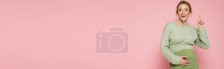 Photo for Positive pregnant woman in sweater pointing with finger isolated on pink, banner - Royalty Free Image