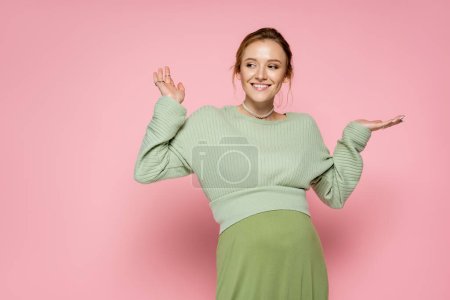 Positiv schwangere Frau im trendigen grünen Outfit zeigt mit Händen auf rosa Hintergrund 
