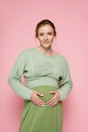 Portrait of positive pregnant woman in green outfit looking at camera isolated on pink 