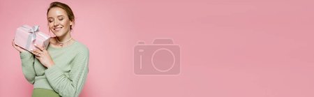 Téléchargez les photos : Joyeux femme enceinte en pull vert tenant boîte cadeau isolé sur rose, bannière - en image libre de droit