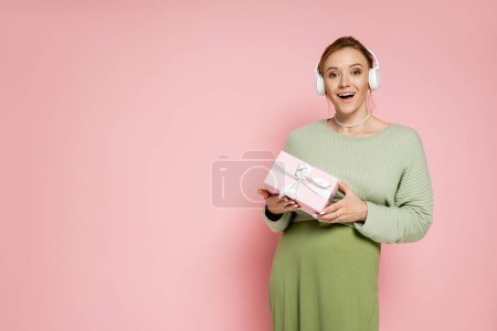 Stylish pregnant woman in headphones holding gift box on pink background 