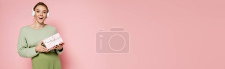 Excited pregnant woman in headphones holding present on pink background, banner 