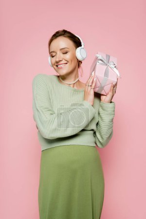 Gut gelaunte Schwangere mit Kopfhörer, Geschenk mit Schleife auf rosa 