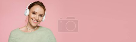 Positive woman in headphones and green sweater looking at camera isolated on pink, banner