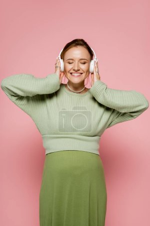 Cheerful pregnant woman in sweater listening music in headphones on pink background 