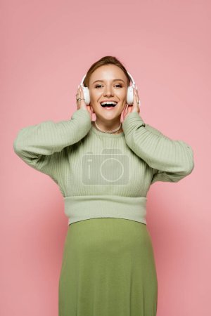 Téléchargez les photos : Femme enceinte excitée écoutant de la musique dans les écouteurs et regardant la caméra sur fond rose - en image libre de droit