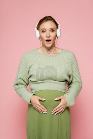 Shocked pregnant woman in green outfit touching belly and using headphones isolated on pink 