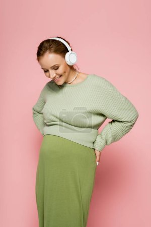 Positive pregnant woman in headphones looking at belly on pink background 