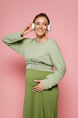 Smiling pregnant woman listening music in headphones and touching belly on pink background 