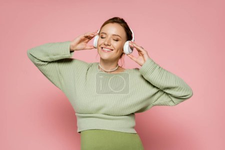 Positive pregnant woman closing eyes while listening music in headphones on pink background 
