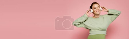 Cheerful pregnant woman in green outfit listening music in headphones on pink background, banner 