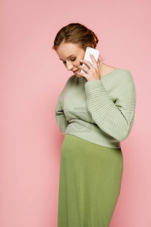 Pretty pregnant woman in green outfit talking on smartphone isolated on pink 