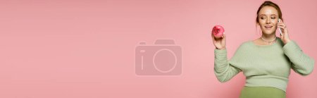 Happy pregnant woman talking on smartphone and looking at sweet donut isolated on pink, banner 
