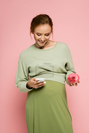Pregnant woman in green outfit using smartphone and holding donut isolated on pink 