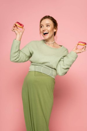 Téléchargez les photos : Femme enceinte excitée en pull tenant des beignets sucrés sur fond rose - en image libre de droit