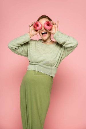 Aufgeregt schwangere Frau bedeckt Gesicht mit Donuts auf rosa Hintergrund 