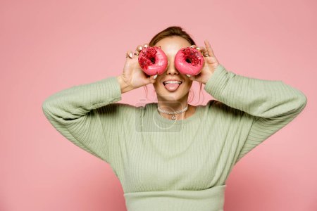 Junge Frau streckt Zunge heraus und hält Donuts in Augennähe isoliert auf rosa 
