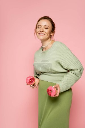 Téléchargez les photos : Femme enceinte souriante tenant de savoureux beignets isolés sur rose - en image libre de droit