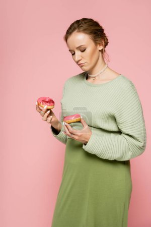 Mujer embarazada pensativa sosteniendo rosquillas dulces aisladas en rosa 
