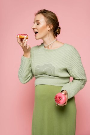 Pregnant woman in green sweater holding donuts isolated on pink 