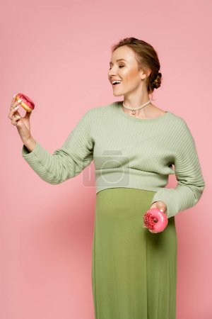 Fröhliche junge Frau in grünem Outfit blickt auf Donut auf rosa Hintergrund 