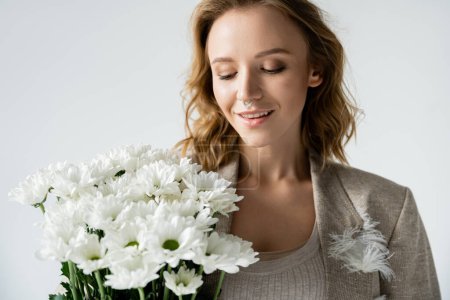Stylish woman in jacket looking at bouquet of white flowers isolated on grey 