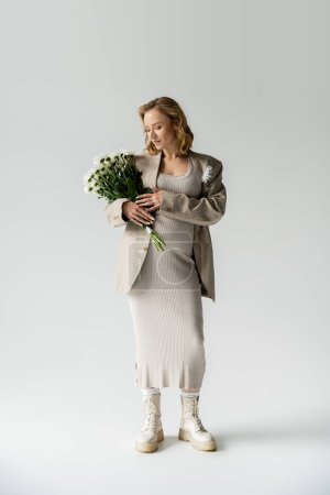 Full length of trendy pregnant woman in dress and jacket holding flowers on grey background 