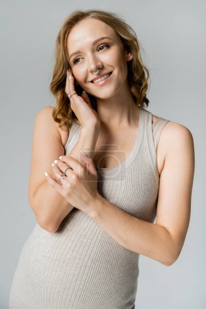 Smiling pregnant woman in dress touching face isolated on grey 