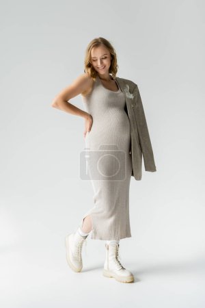 Stilvolle schwangere Frau in Kleid und Stiefeln posiert auf grauem Hintergrund 