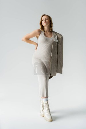Trendige schwangere Frau in Kleid und Jacke posiert auf grauem Hintergrund 