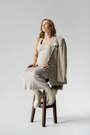 Fashionable pregnant woman in dress and jacket sitting on chair on grey background 