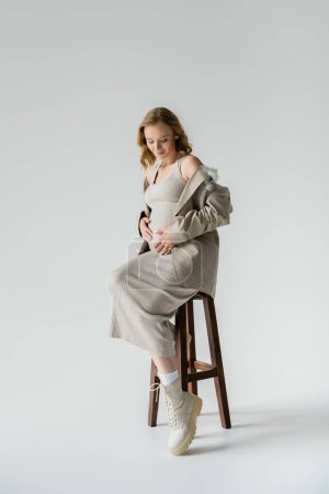 Full length of positive pregnant woman in dress sitting on chair on grey background 
