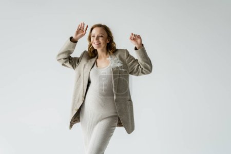 Photo for Cheerful pregnant woman in dress and jacket waving hand isolated on grey - Royalty Free Image
