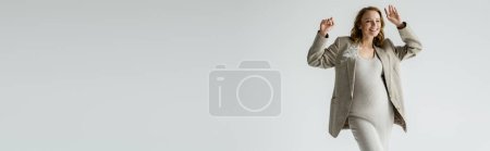 Positive pregnant woman in stylish jacket and dress waving hand isolated on grey, banner 