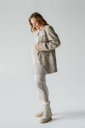 Full length of stylish pregnant woman in dress looking at belly on grey background 