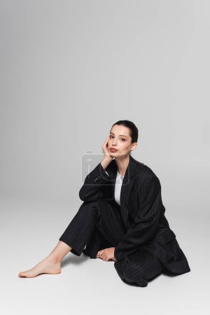 Téléchargez les photos : Modèle élégant pieds nus en costume assis sur fond gris - en image libre de droit