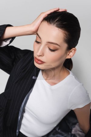 High angle view of brunette model in t-shirt and jacket touching hair isolated on grey 