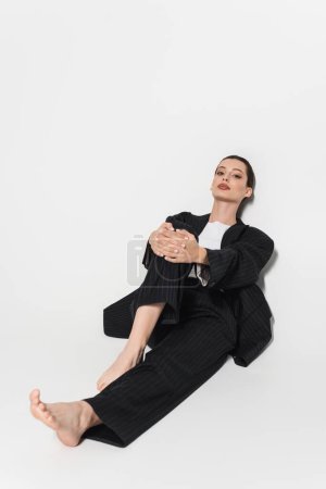 Full length of trendy model in suit touching knee on white background 