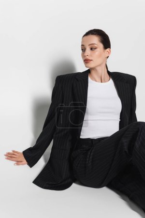 Téléchargez les photos : Femme brune élégante en costume rayé assis sur fond blanc avec ombre - en image libre de droit