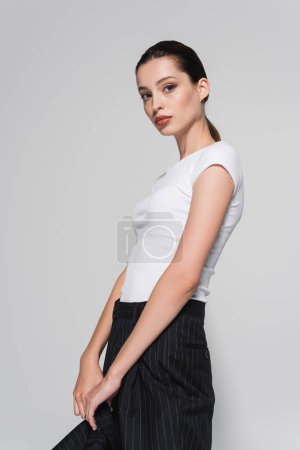 Stylish woman in white t-shirt holding jacket ad looking at camera isolated on grey 