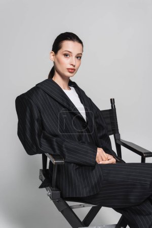 Stilvolle Frau im Anzug sitzt auf Klappstuhl auf grauem Hintergrund 